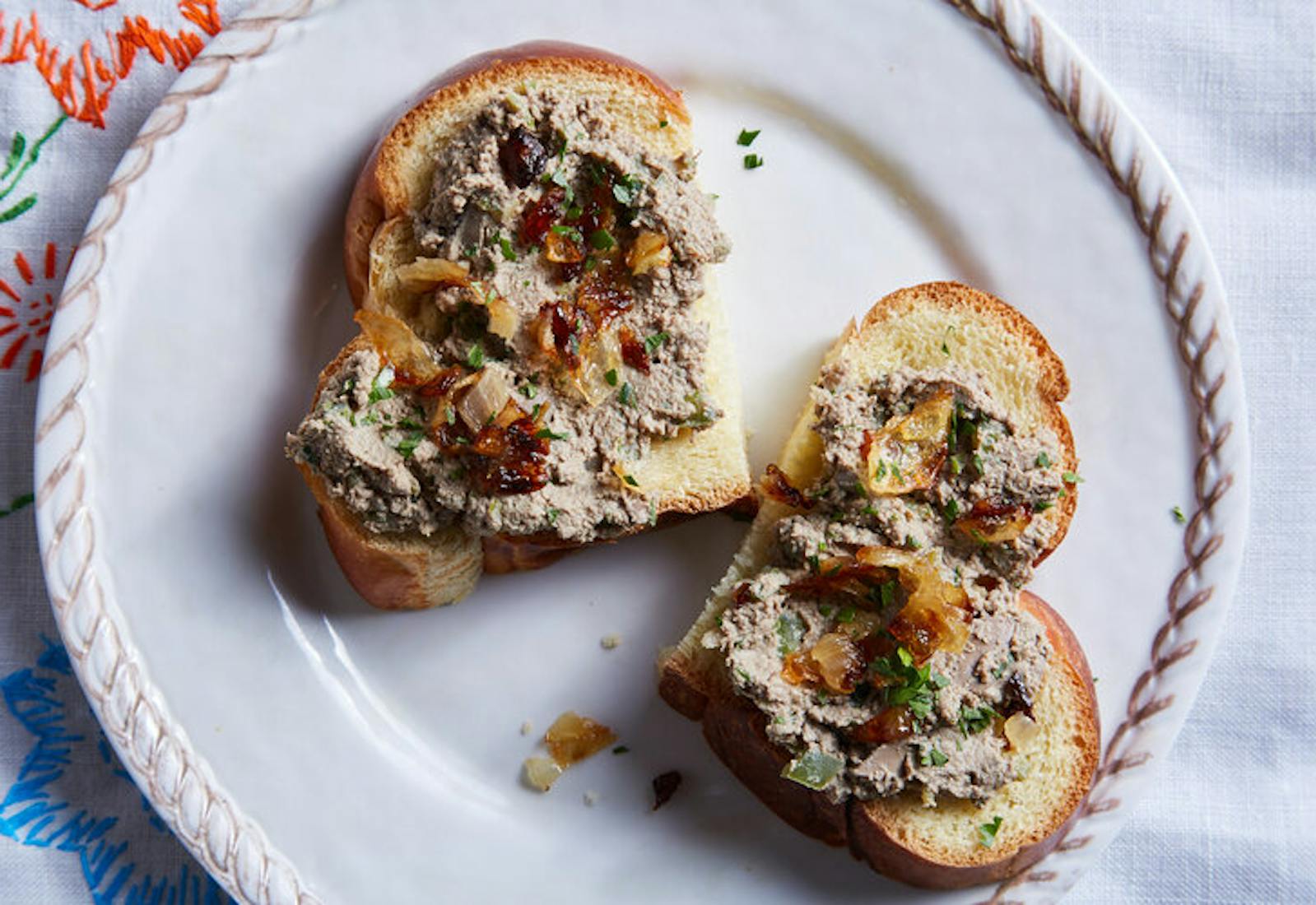 Chopped liver with caramelized onions and parsley on sliced challah, atop embroidered white tablecloth.