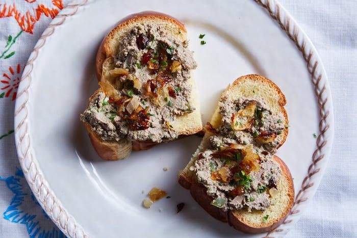 Chopped liver with caramelized onions and parsley on sliced challah, atop embroidered white tablecloth.