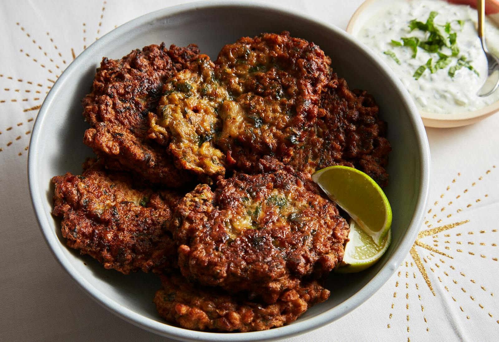 Cauliflower beignets with lime wedges and yogurt sauce atop white tablecloth with star patterns.