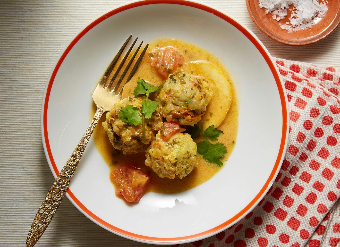Fish balls in saffron tomato sauce garnished with sprigs of cilantro on white plate with red polka-dot napkin, atop light cream tablecloth.