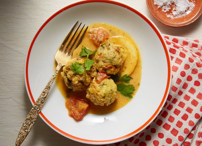 Fish balls in saffron tomato sauce garnished with sprigs of cilantro on white plate with red polka-dot napkin, atop light cream tablecloth.