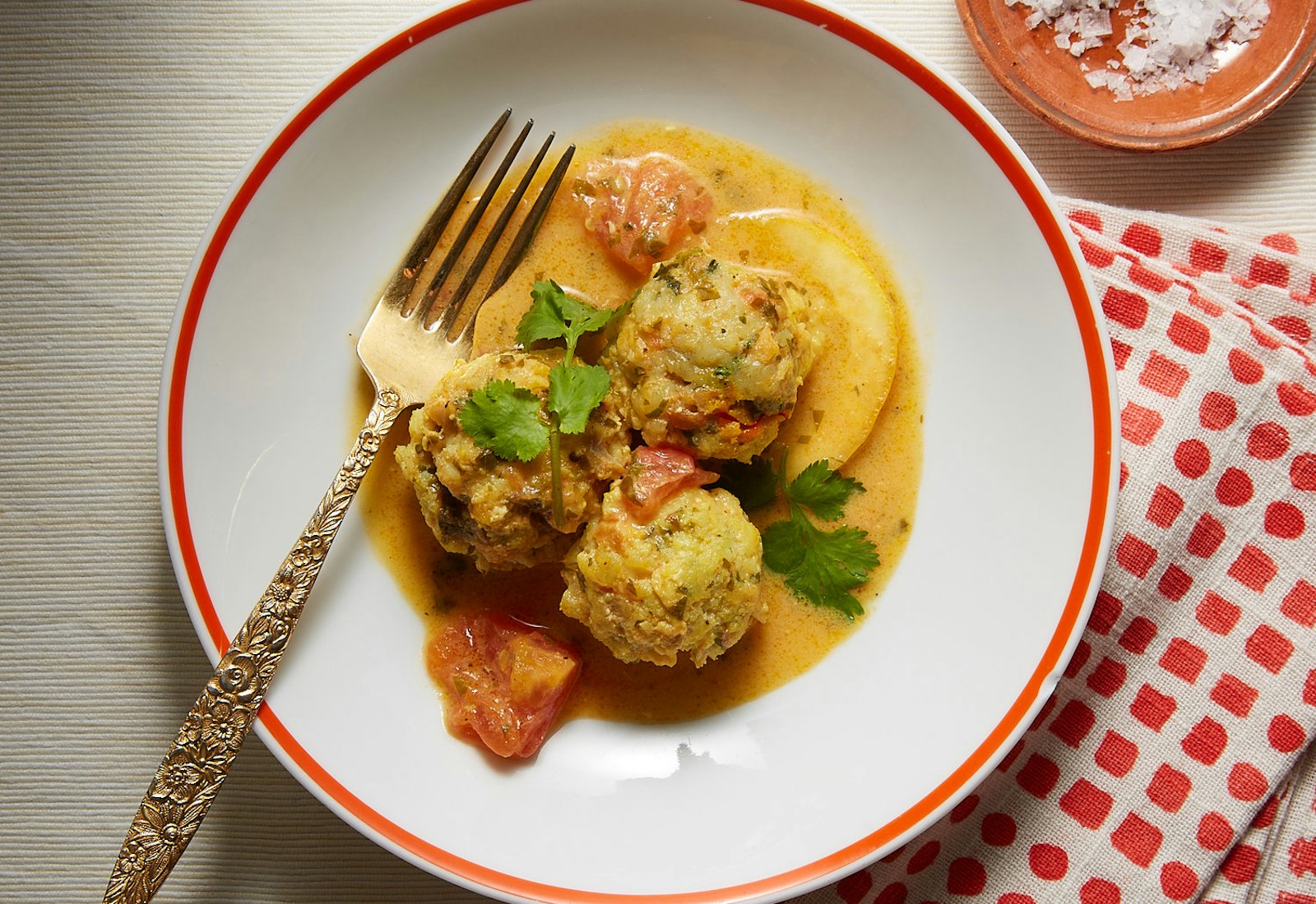 Fish balls in saffron tomato sauce garnished with sprigs of cilantro on white plate with red polka-dot napkin, atop light cream tablecloth.