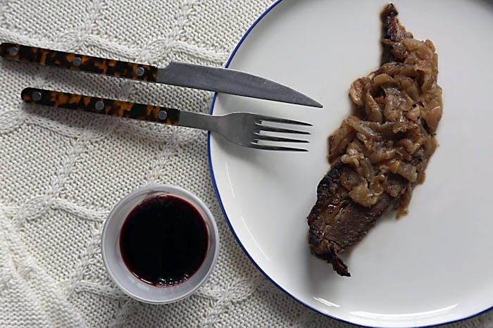 Slice of brisket with onions on white plate alongside cup of red wine atop white woven tablecloth.