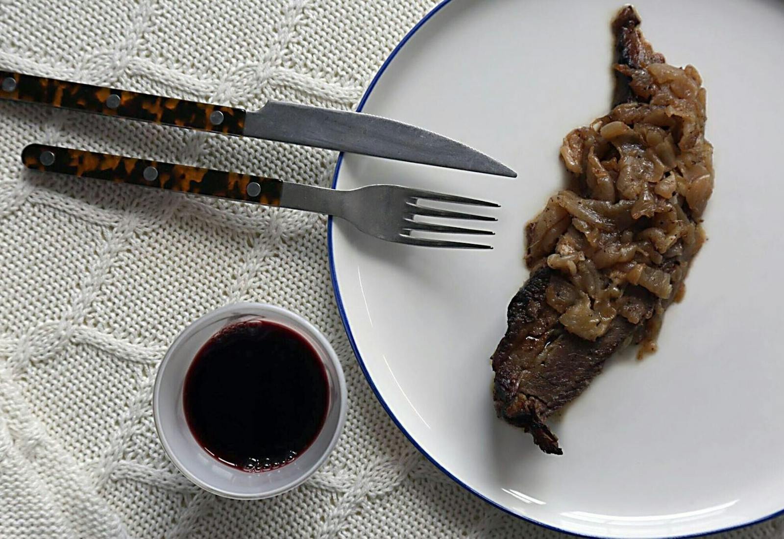 Slice of brisket with onions on white plate alongside cup of red wine atop white woven tablecloth.