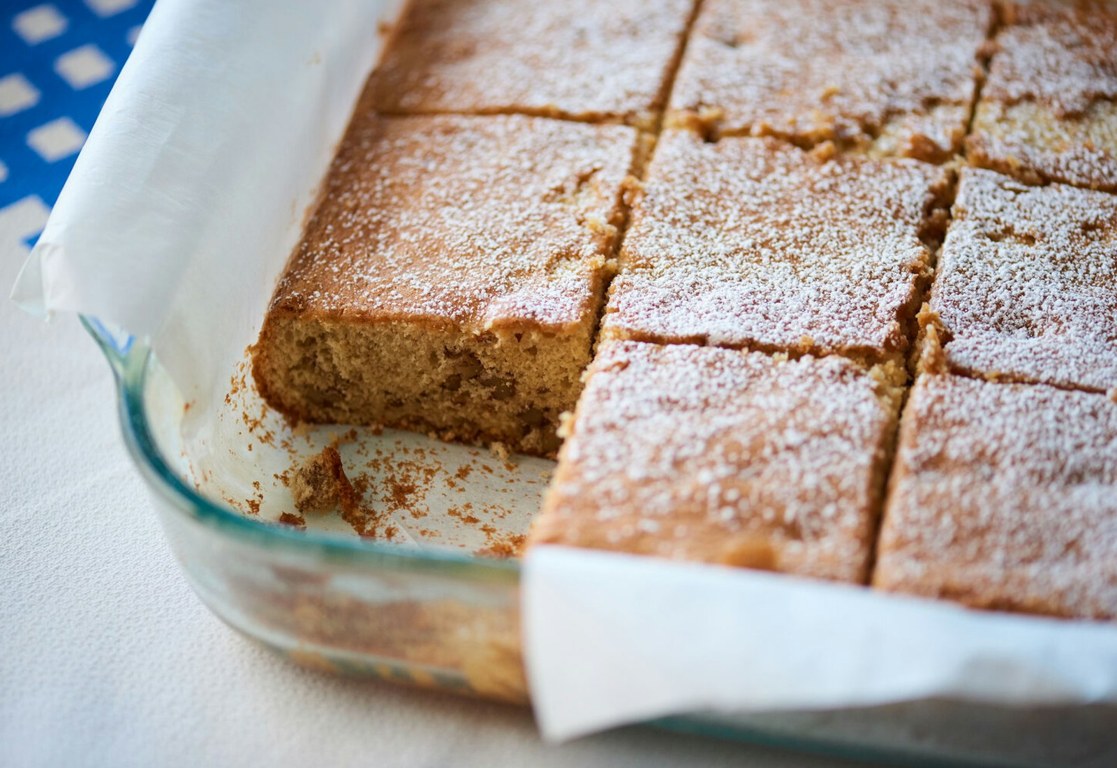 Sliced maple walnut cake in parchment lined baking dish, atop blue and white checkered tablecloth.