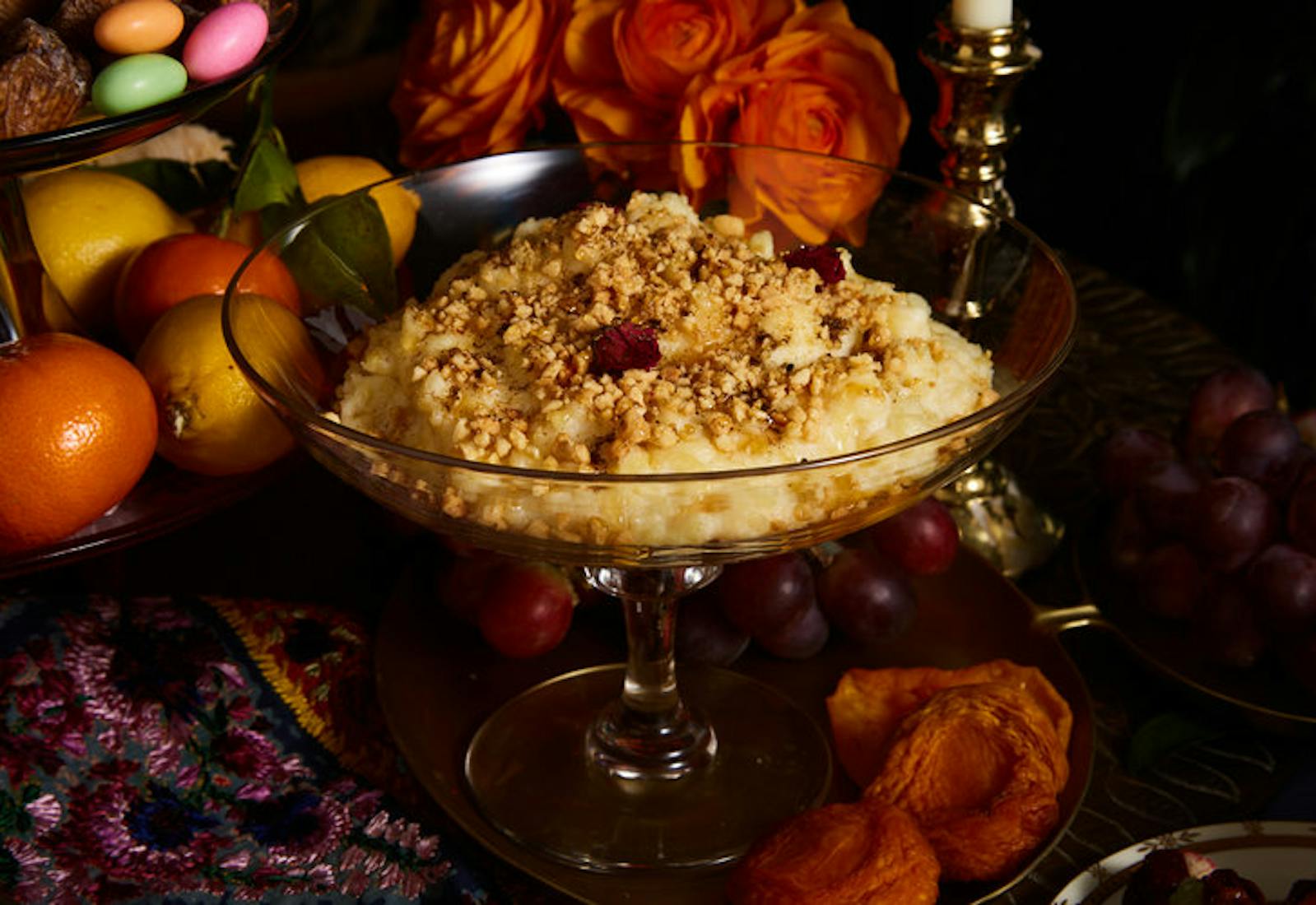 Sweet couscous topped with nuts and dried fruit in glass chalice, surrounded by fresh and dry fruits.