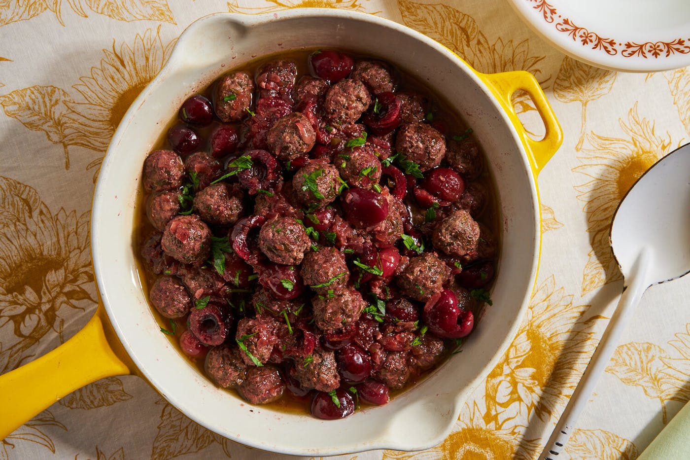 Meatballs and cherries with sliced mint leaves in ceramic dish atop yellow floral tablecloth.