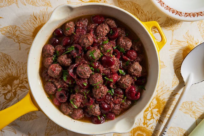 Meatballs and cherries with sliced mint leaves in ceramic dish atop yellow floral tablecloth.