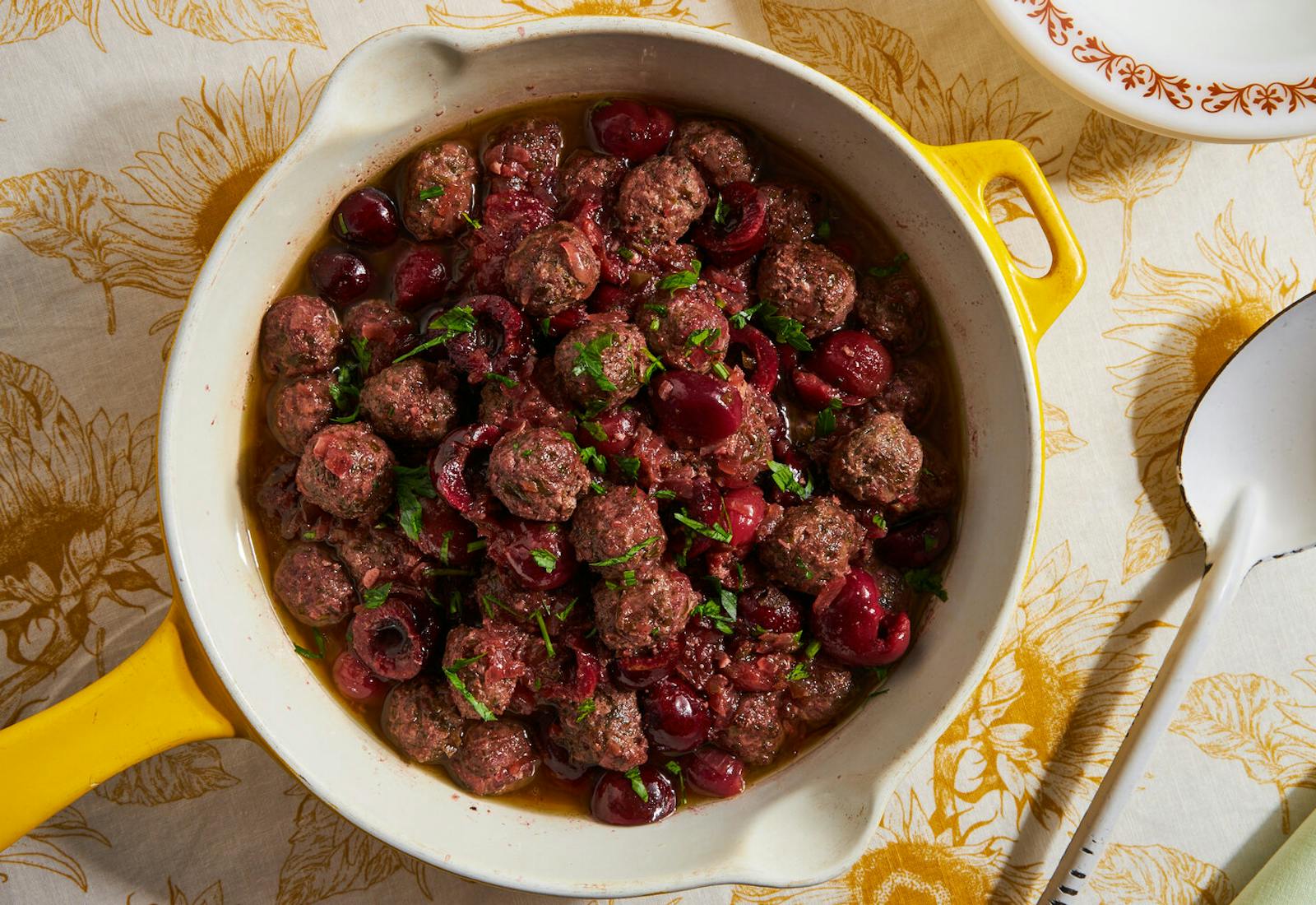 Meatballs and cherries with sliced mint leaves in ceramic dish atop yellow floral tablecloth.