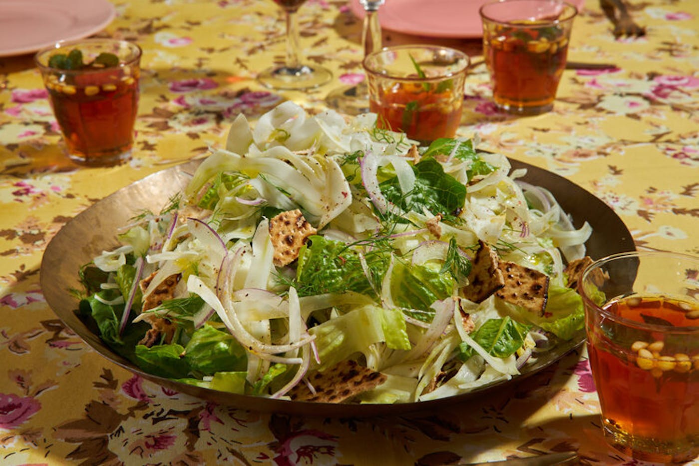 Lettuce and fennel salad garnished with pieces of matzo, mint tea atop yellow floral tablecloth.