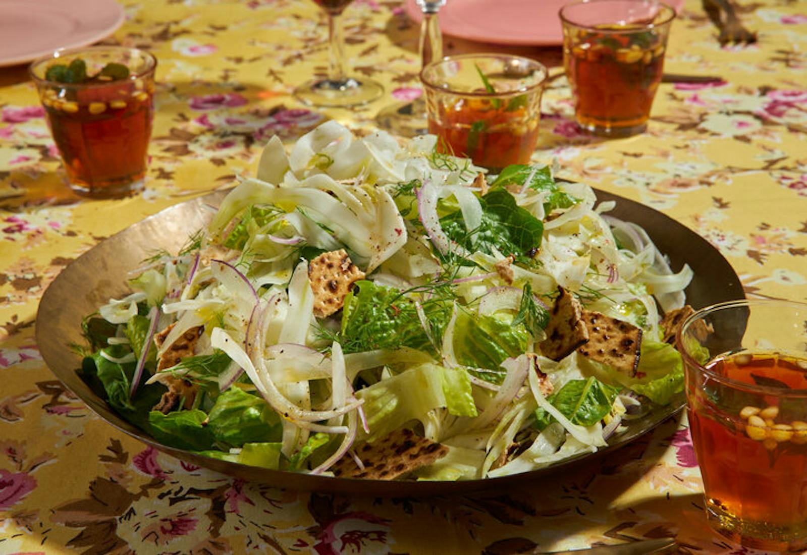 Lettuce and fennel salad garnished with pieces of matzo, mint tea atop yellow floral tablecloth.