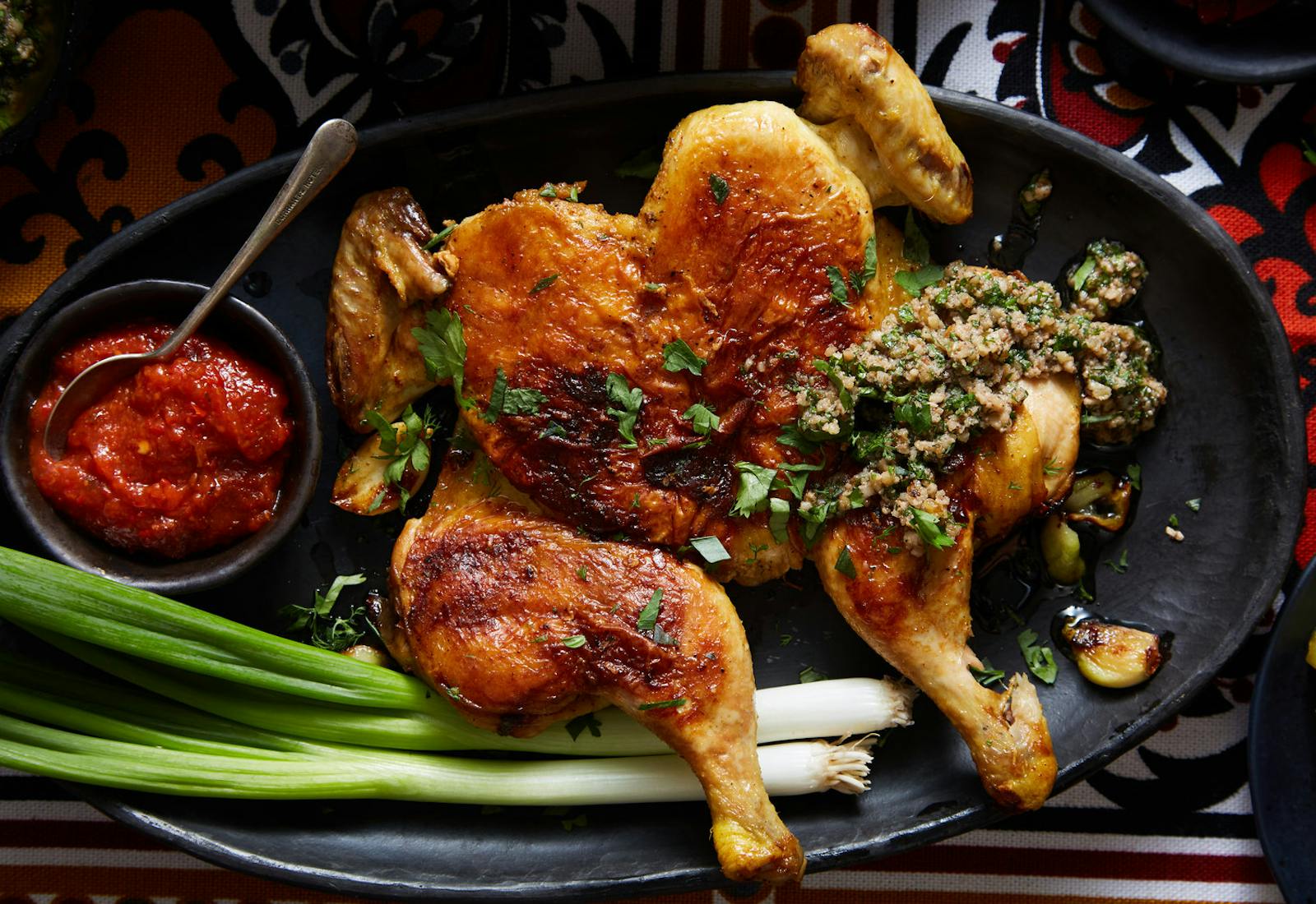 Pan fried cornish hen with radishes,  walnut garlic sauce and adjika sauce on black plate atop red and orange tablecloth.