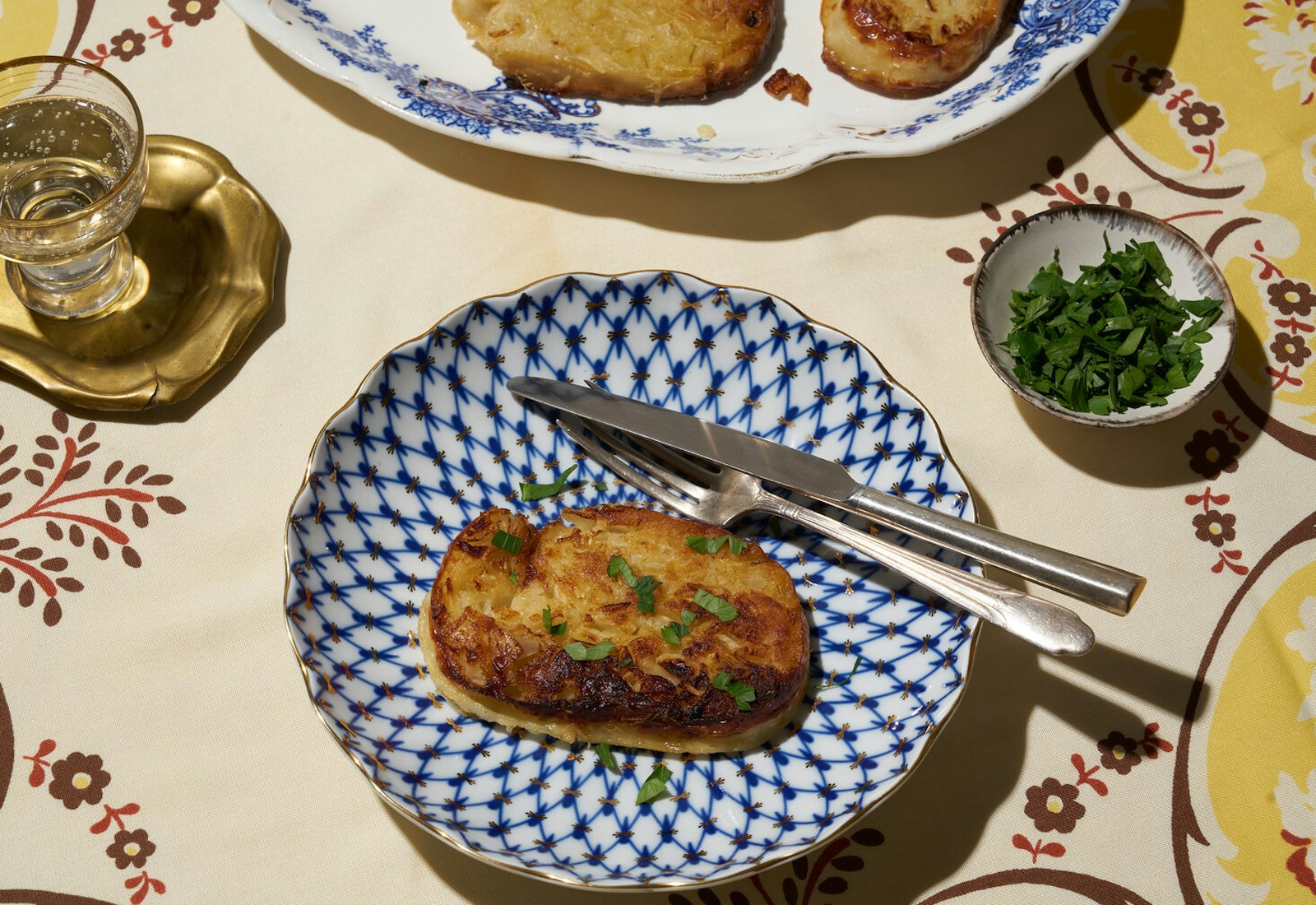 Stuffed chicken neck with chopped parsley alongside glass of vodka and dish of fresh parsley atop yellow floral tablecloth.