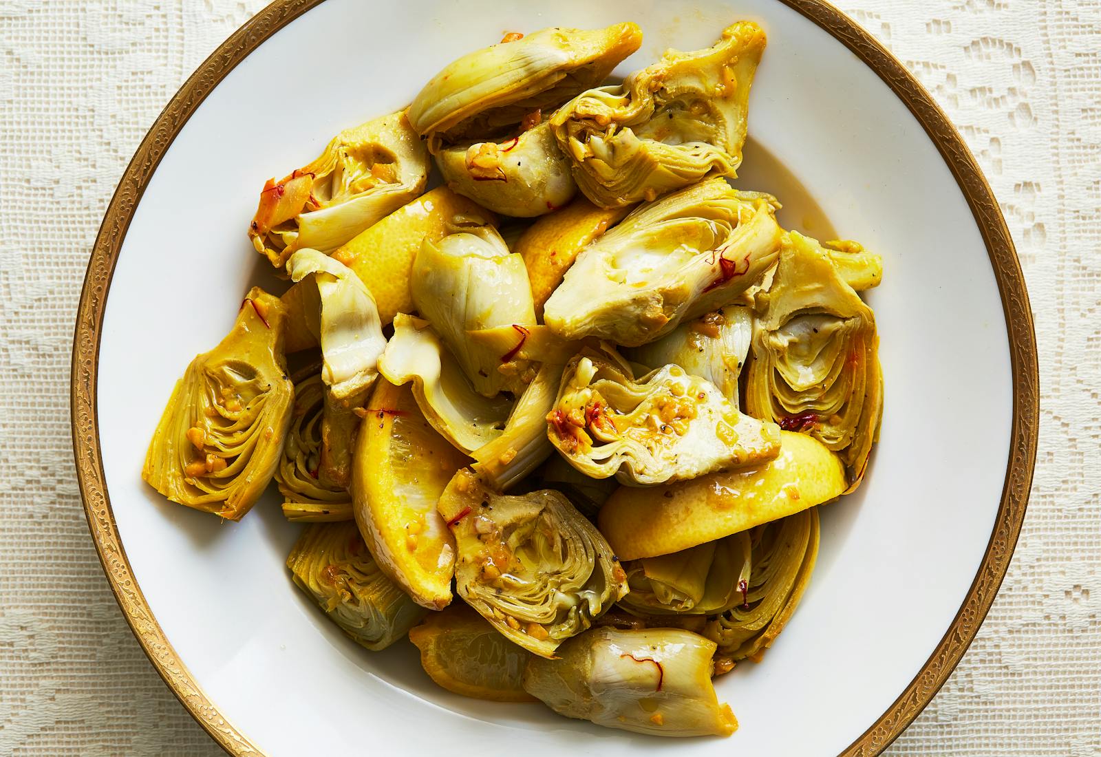Sliced artichoke hearts and lemon slices in white bowl with golden rim.