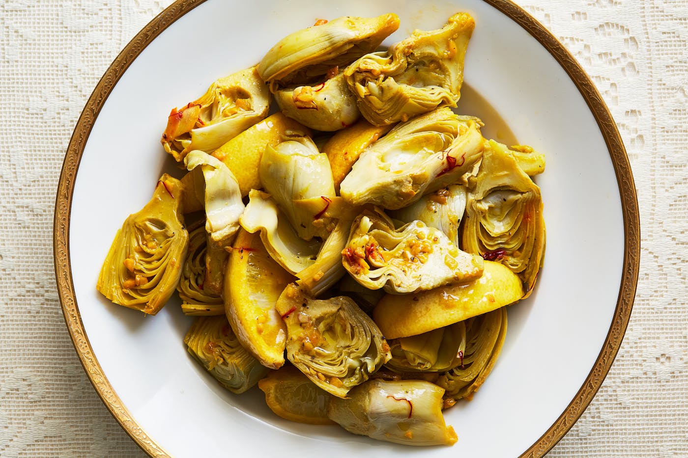 Sliced artichoke hearts and lemon slices in white bowl with golden rim.