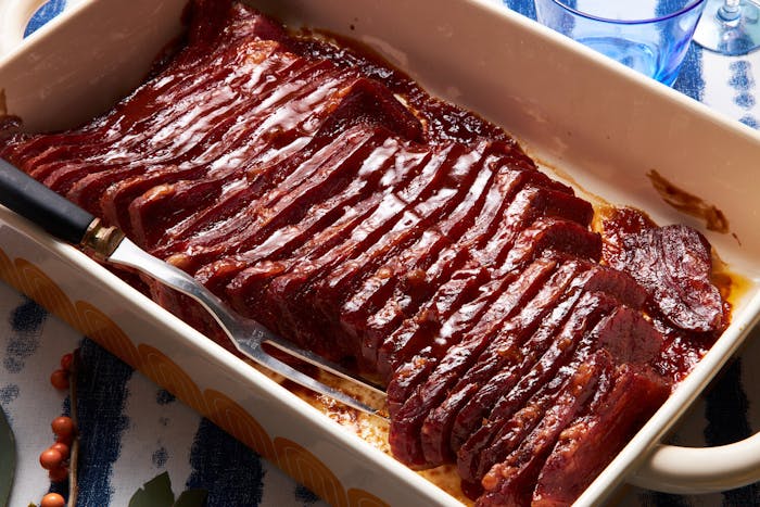 Sliced corned beef in white casserole pan, serving fork atop blue tablecloth.