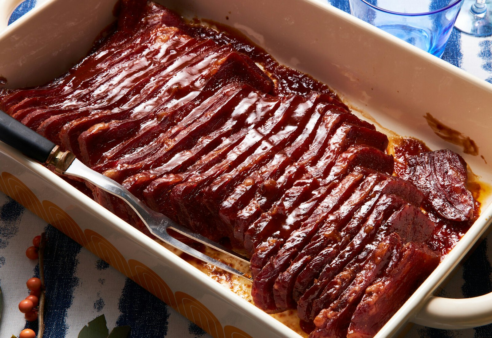 Sliced corned beef in white casserole pan, serving fork atop blue tablecloth.