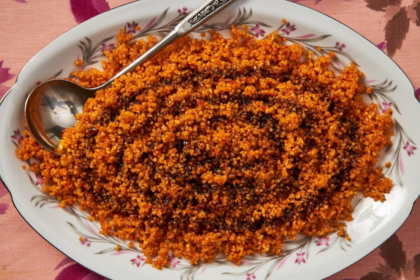 Bulgur muhammara with drizzle of pomegranate molasses in large serving dish atop pink tablecloth.