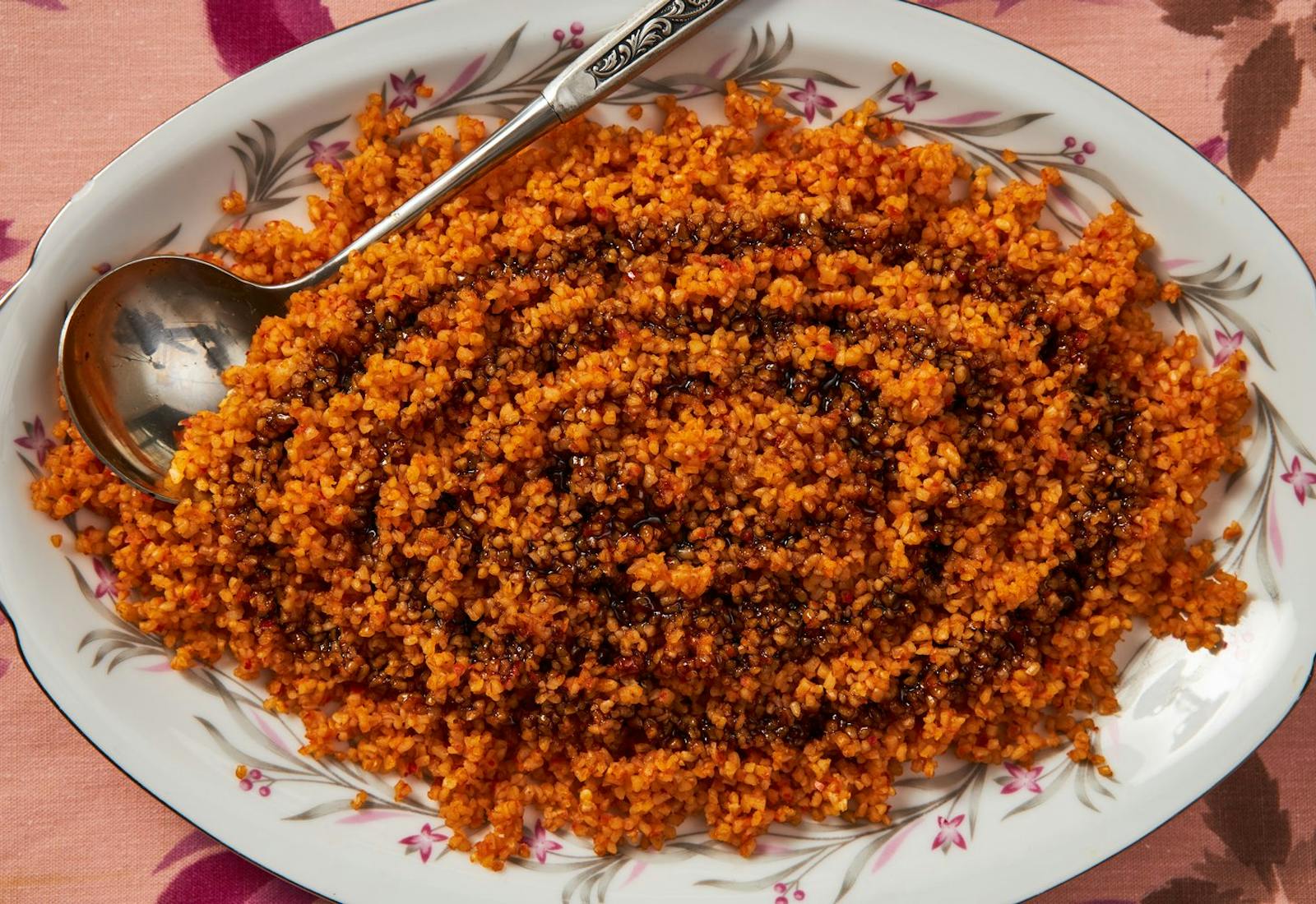 Bulgur muhammara with drizzle of pomegranate molasses in large serving dish atop pink tablecloth.