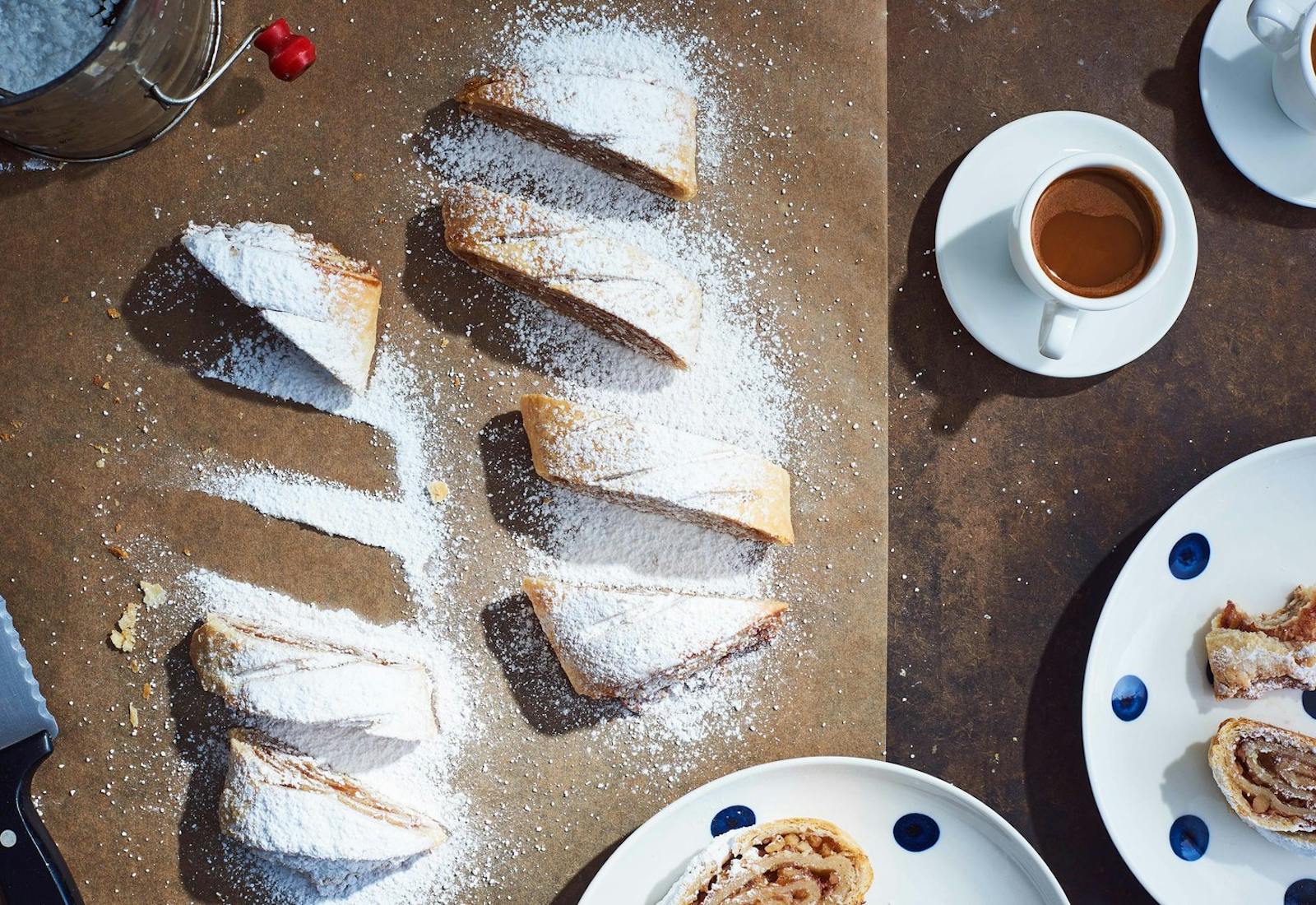 Sliced apricot walnut strudel generously coated in powdered sugar alongside polkadot dishes of strudel, mugs of coffee and a sifter with powdered sugar, atop parchment paper and brown table.