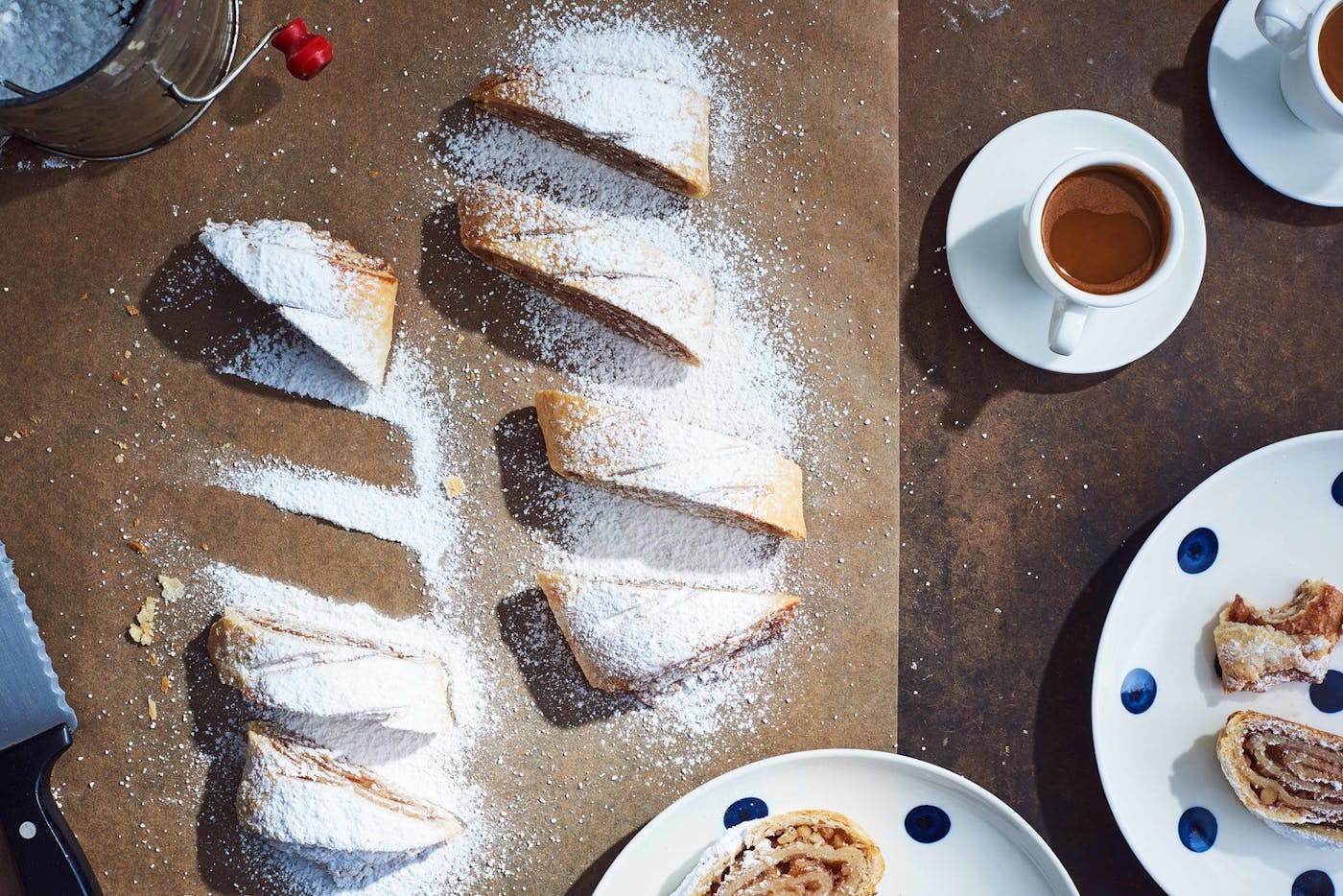 Sliced apricot walnut strudel generously coated in powdered sugar alongside polkadot dishes of strudel, mugs of coffee and a sifter with powdered sugar, atop parchment paper and brown table.