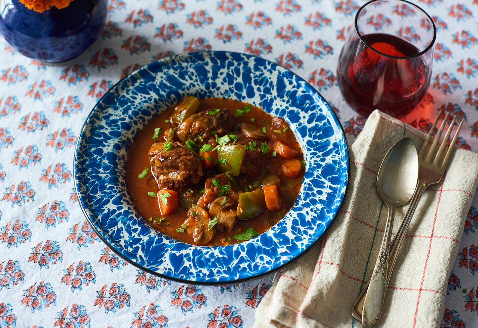 Goulash garnished with parsley in blue speckled bowl, red wine atop floral tablecloth.