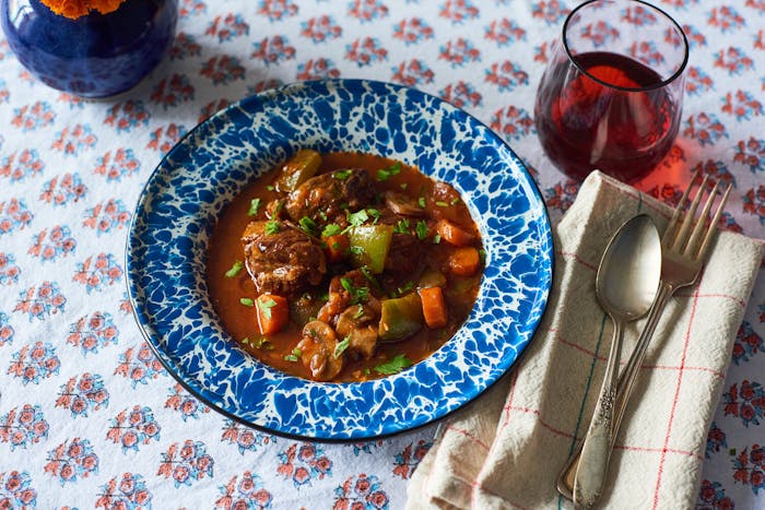 Goulash garnished with parsley in blue speckled bowl, red wine atop floral tablecloth.