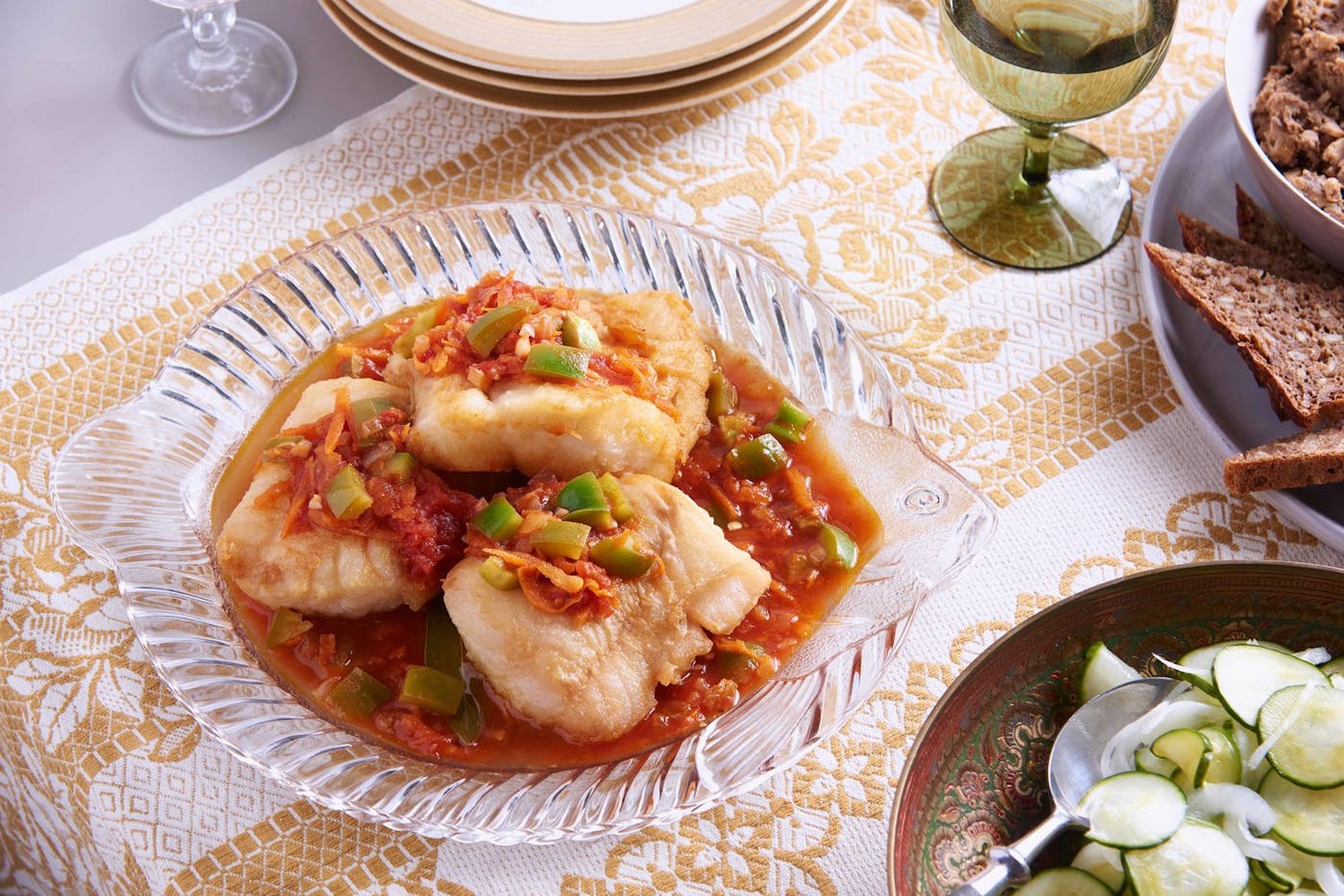 Baked fish in sweet and sour sauce in glass fish-shaped dish, alongside pickled cucumber salad, sliced bread and glass of white wine.