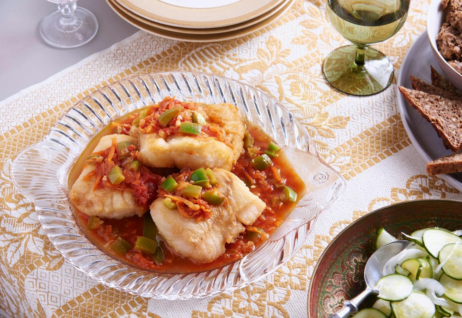 Baked fish in sweet and sour sauce in glass fish-shaped dish, alongside pickled cucumber salad, sliced bread and glass of white wine.