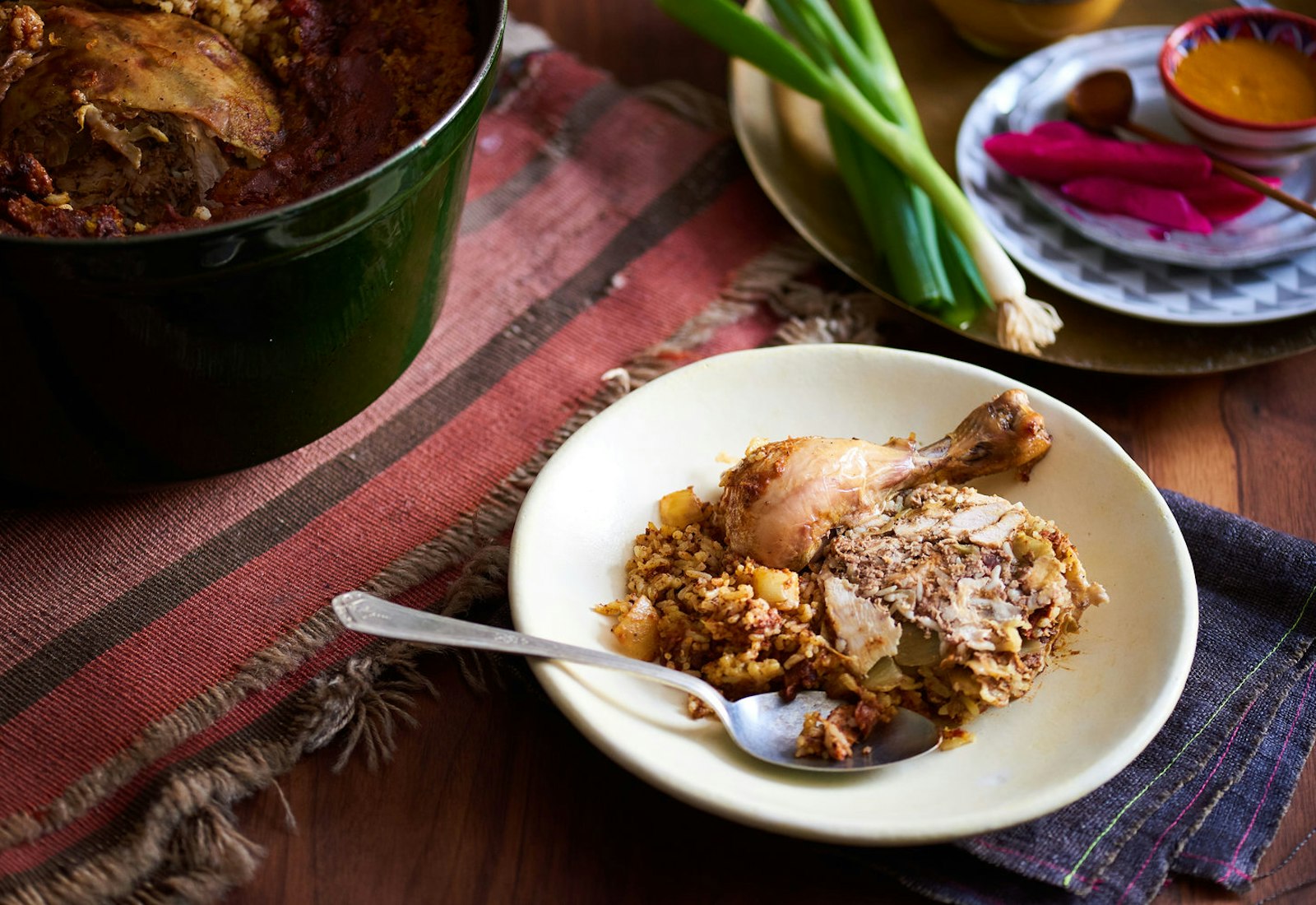 Serving of t'bit alongside large casserole dish of t'bit with plate of scallions and pickled turnips, atop colorful napkins and wooden surface.