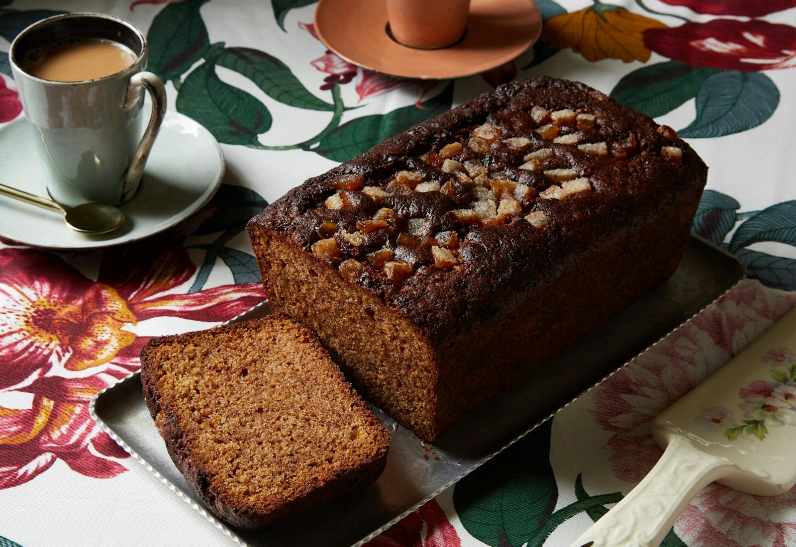 Persimmon loaf on tin plate alongside mugs of coffee, atop floral tablecloth.