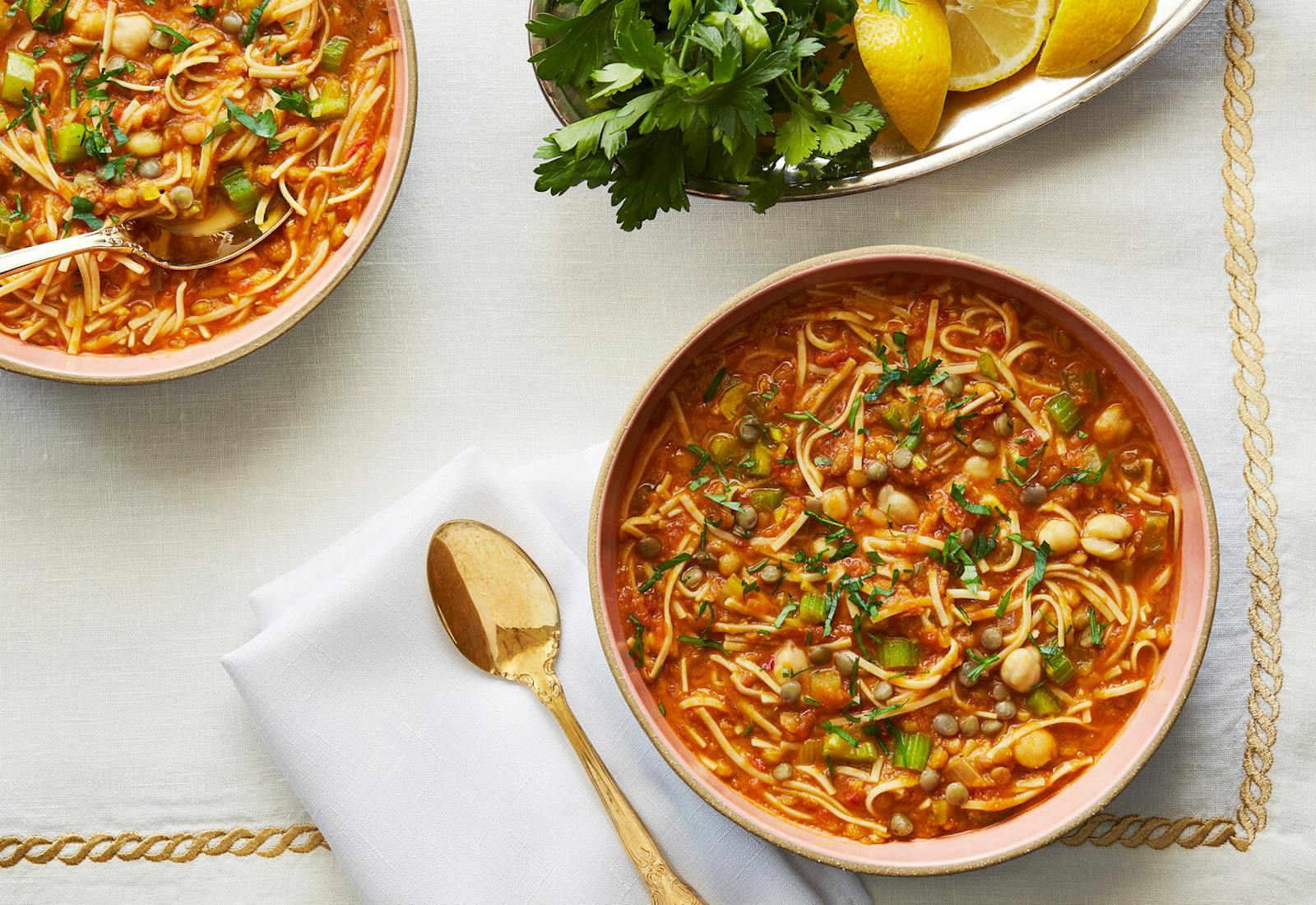 Two bowls of harira alongside dish with parsley and lemon wedges atop white and gold tablecloth.