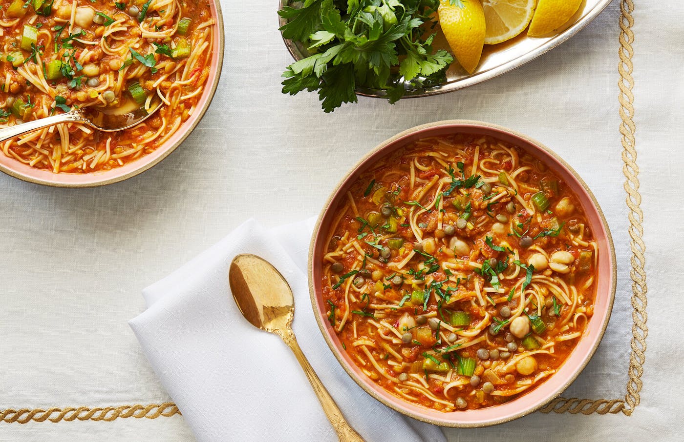 Two bowls of harira alongside dish with parsley and lemon wedges atop white and gold tablecloth.