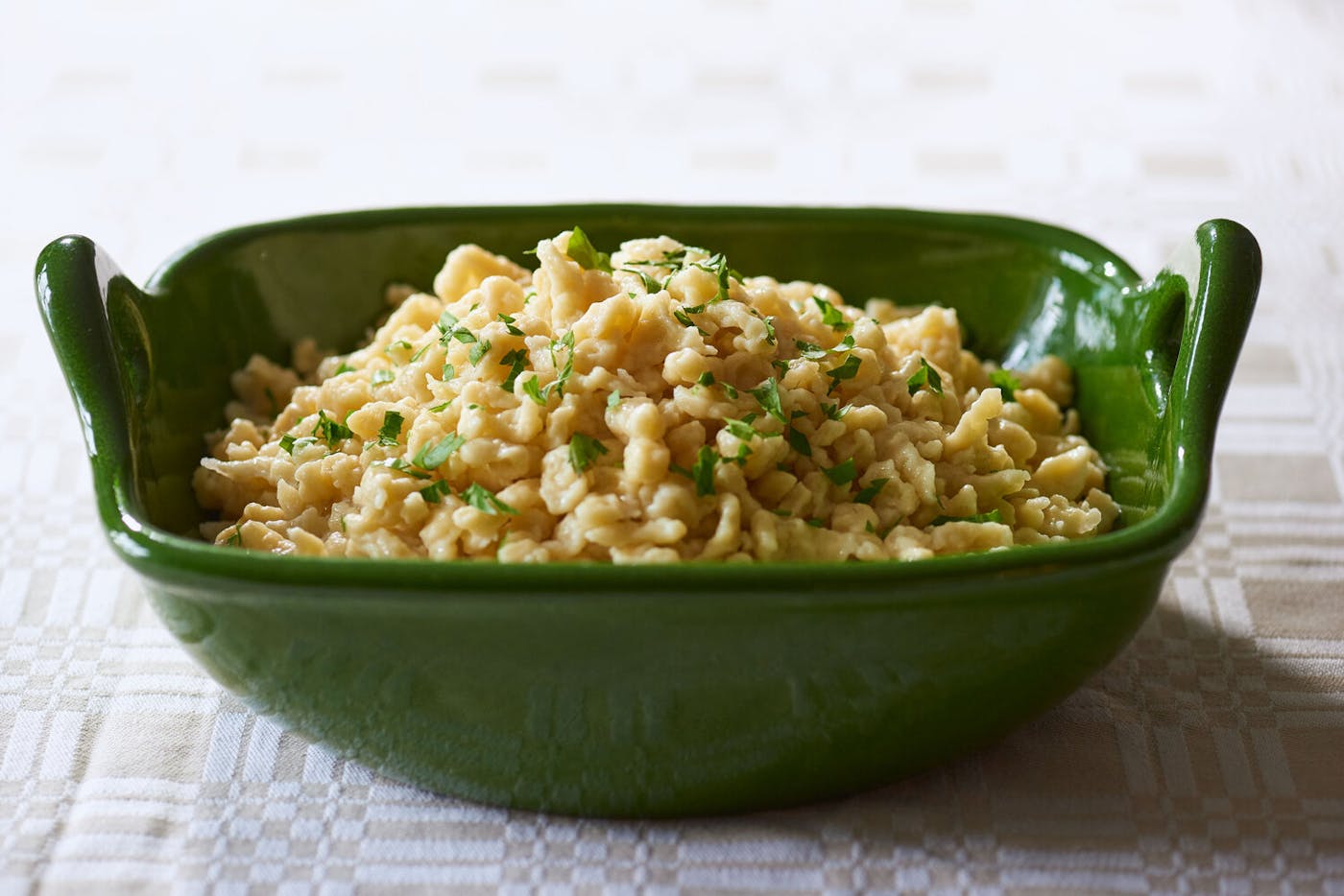 Spaetzle with sliced parsley in green serving bowl, atop tan and white tablecloth.