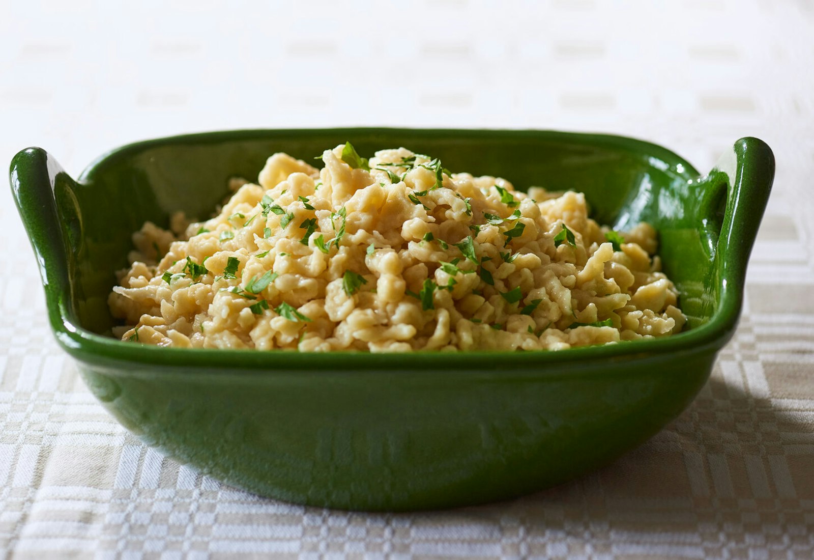 Spaetzle with sliced parsley in green serving bowl, atop tan and white tablecloth.