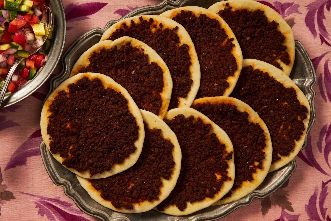 Laham b'Ajeen on a platter, side of chopped salad atop pink floral tablecloth.