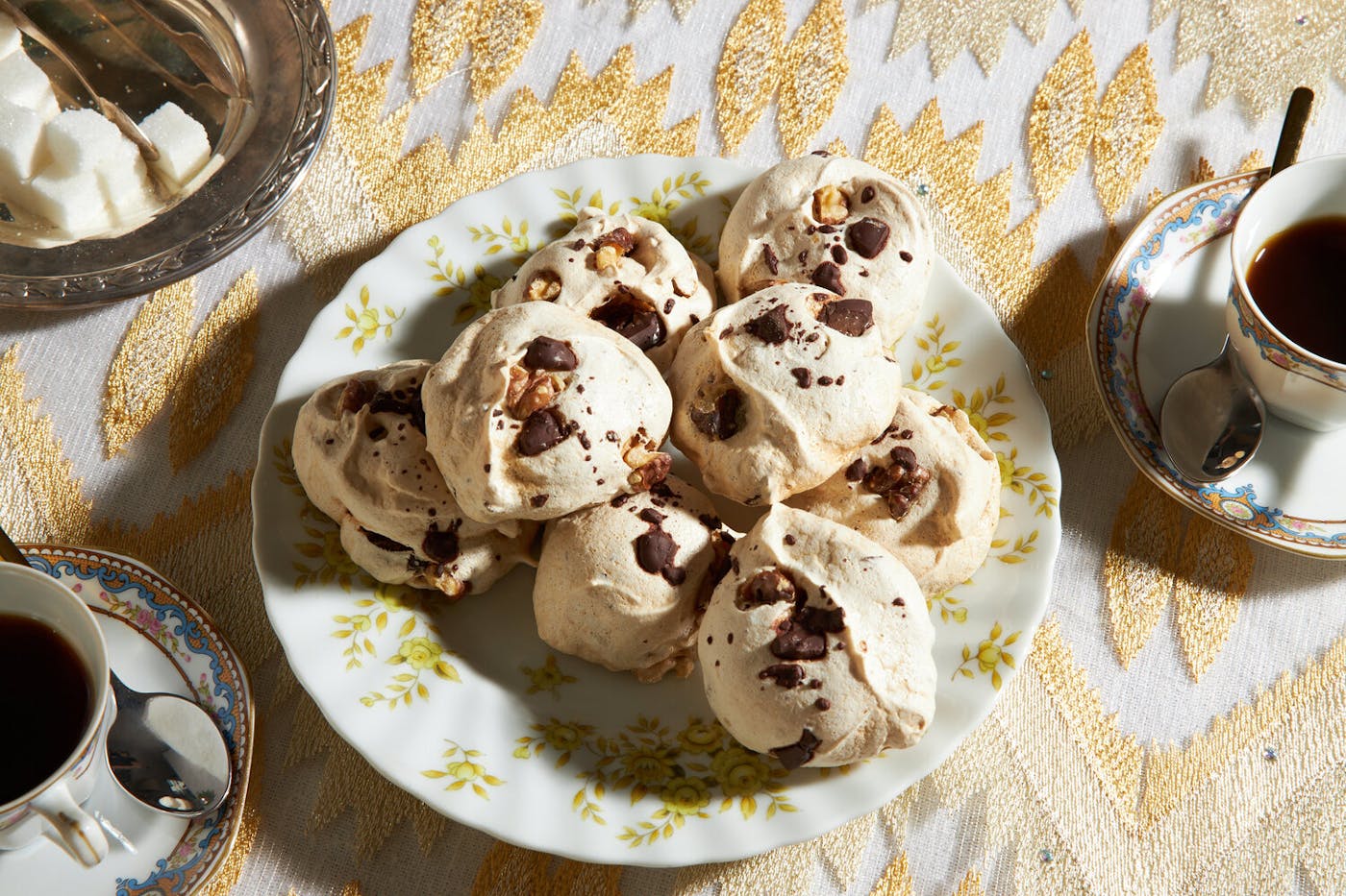 Meringues with sugar cubes and teacups atop gold tablecloth. 