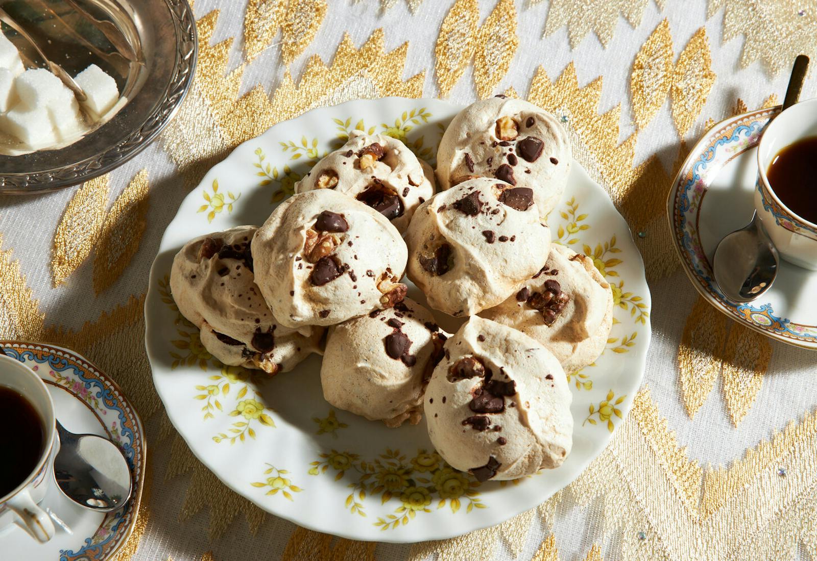 Meringues with sugar cubes and teacups atop gold tablecloth. 