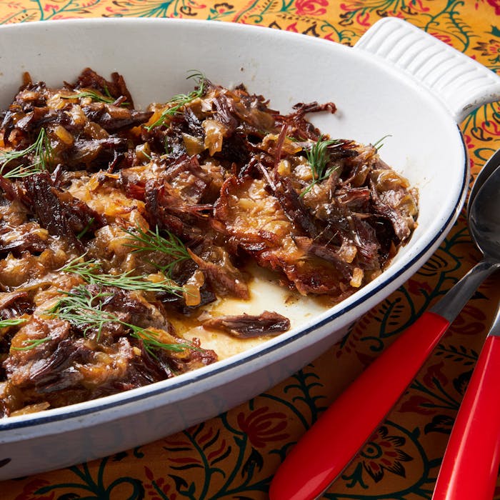Latkes with braised shorts ribs with sprigs of dill in white casserole dish, atop yellow floral tablecloth.