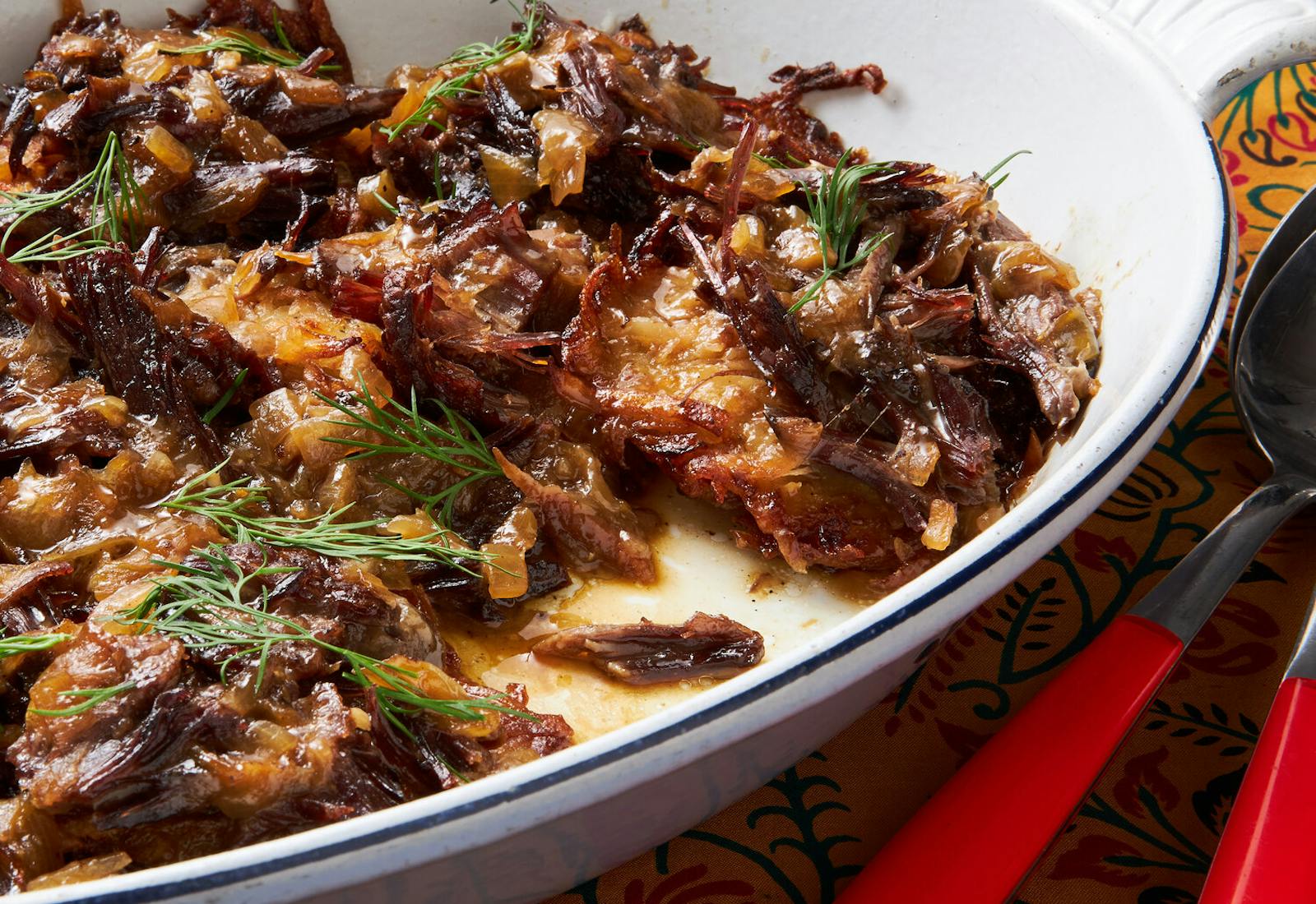 Latkes with braised shorts ribs with sprigs of dill in white casserole dish, atop yellow floral tablecloth.