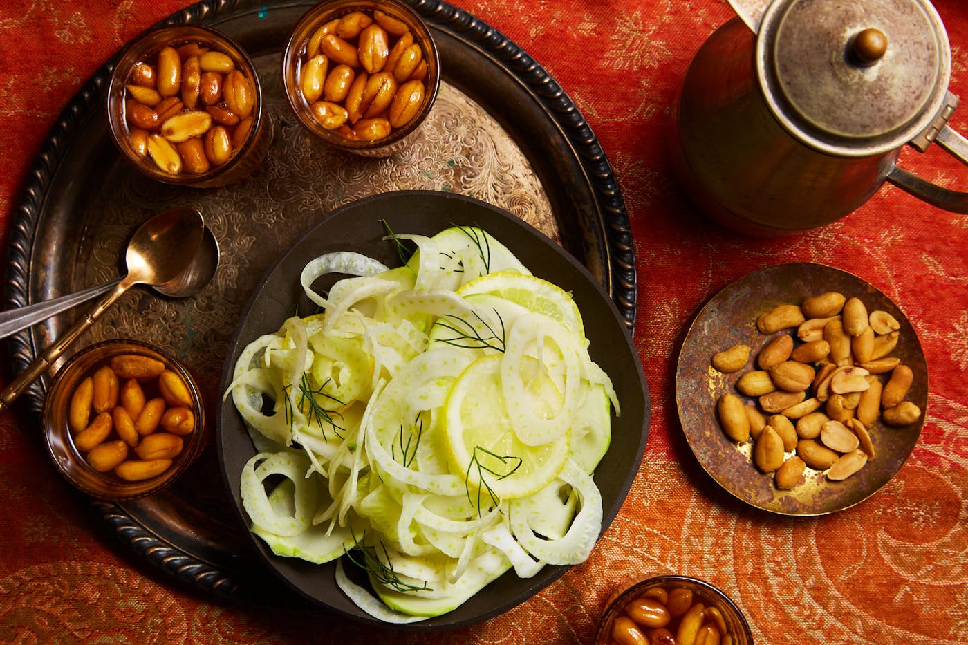 Glasses of Libyan tea with kettle alongside bowls of peanuts and shaved fennel, atop bright red patterned tablecloth.