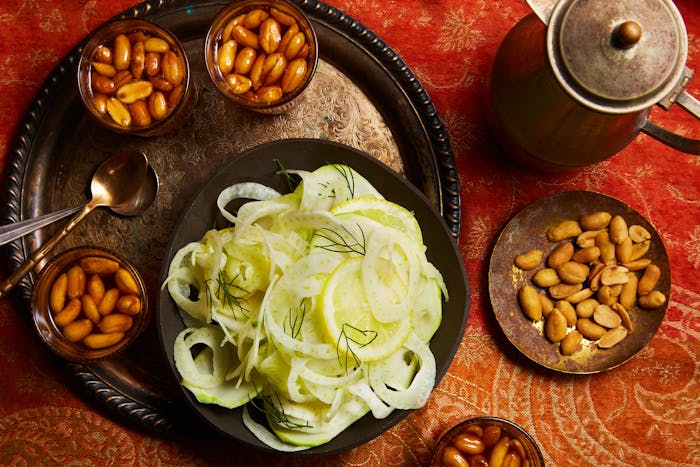 Glasses of Libyan tea with kettle alongside bowls of peanuts and shaved fennel, atop bright red patterned tablecloth.