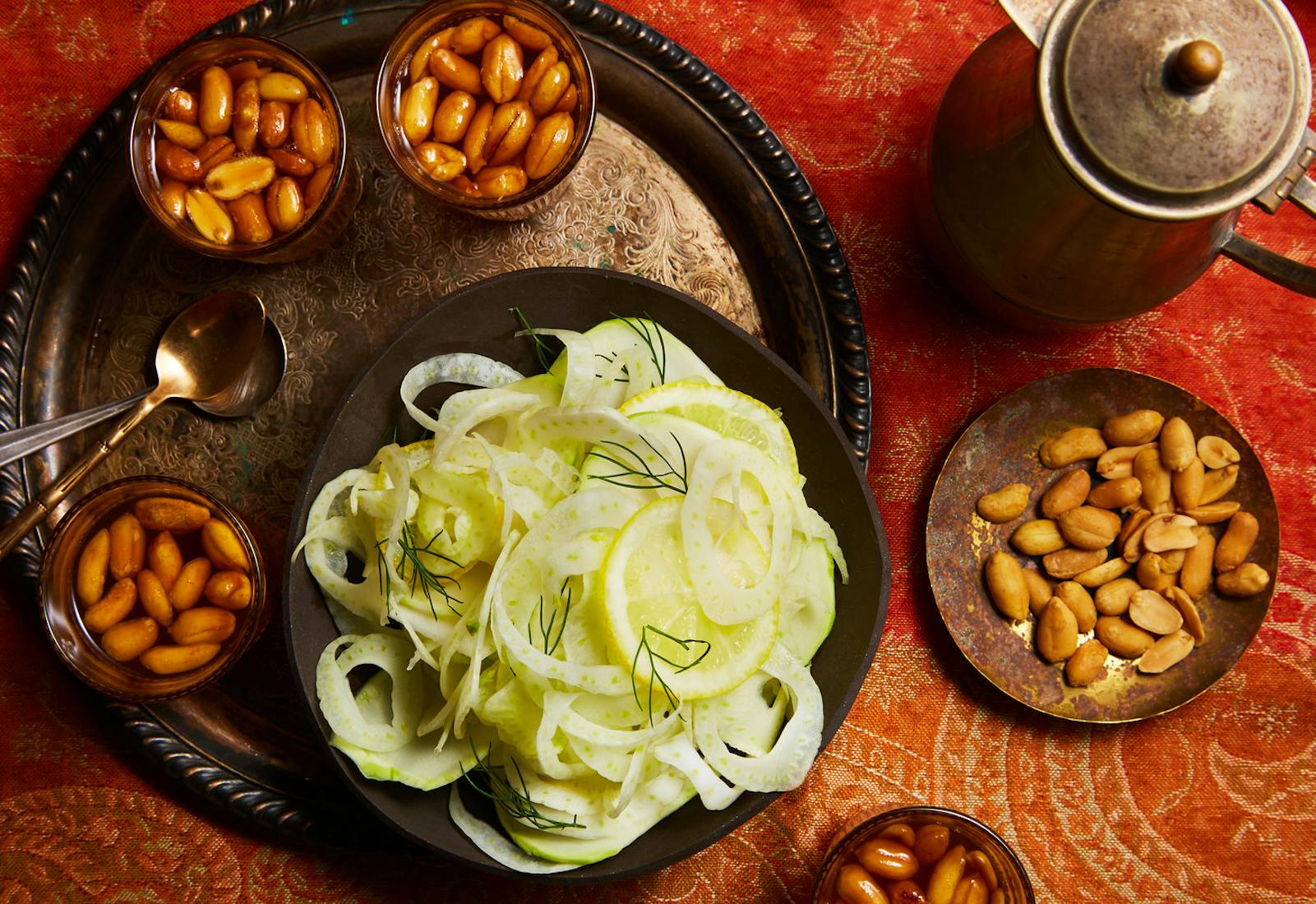 Glasses of Libyan tea with kettle alongside bowls of peanuts and shaved fennel, atop bright red patterned tablecloth.