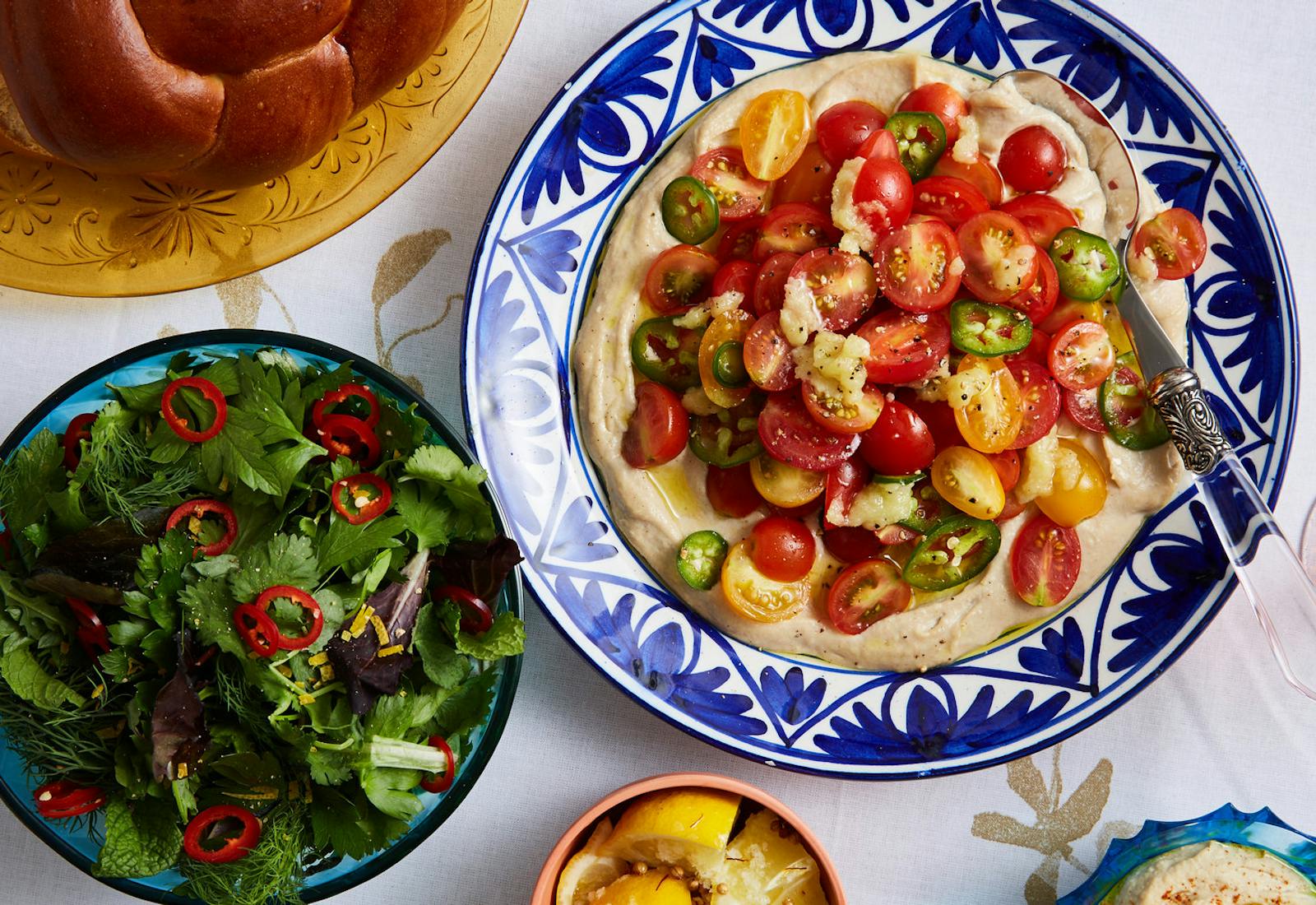 Tomato and garlic salad with tahini dressing in blue bowl alongside herb salad, challah and spiced lemon wedges.