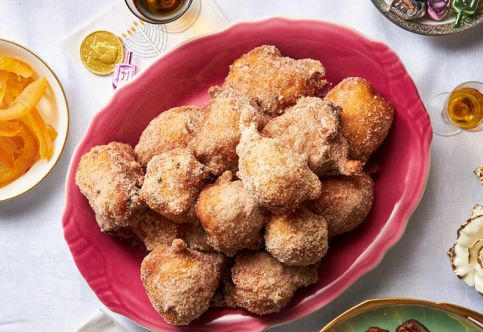 Frittole dolci with gelt, dreidels, wine and flowers atop white tablecloth.