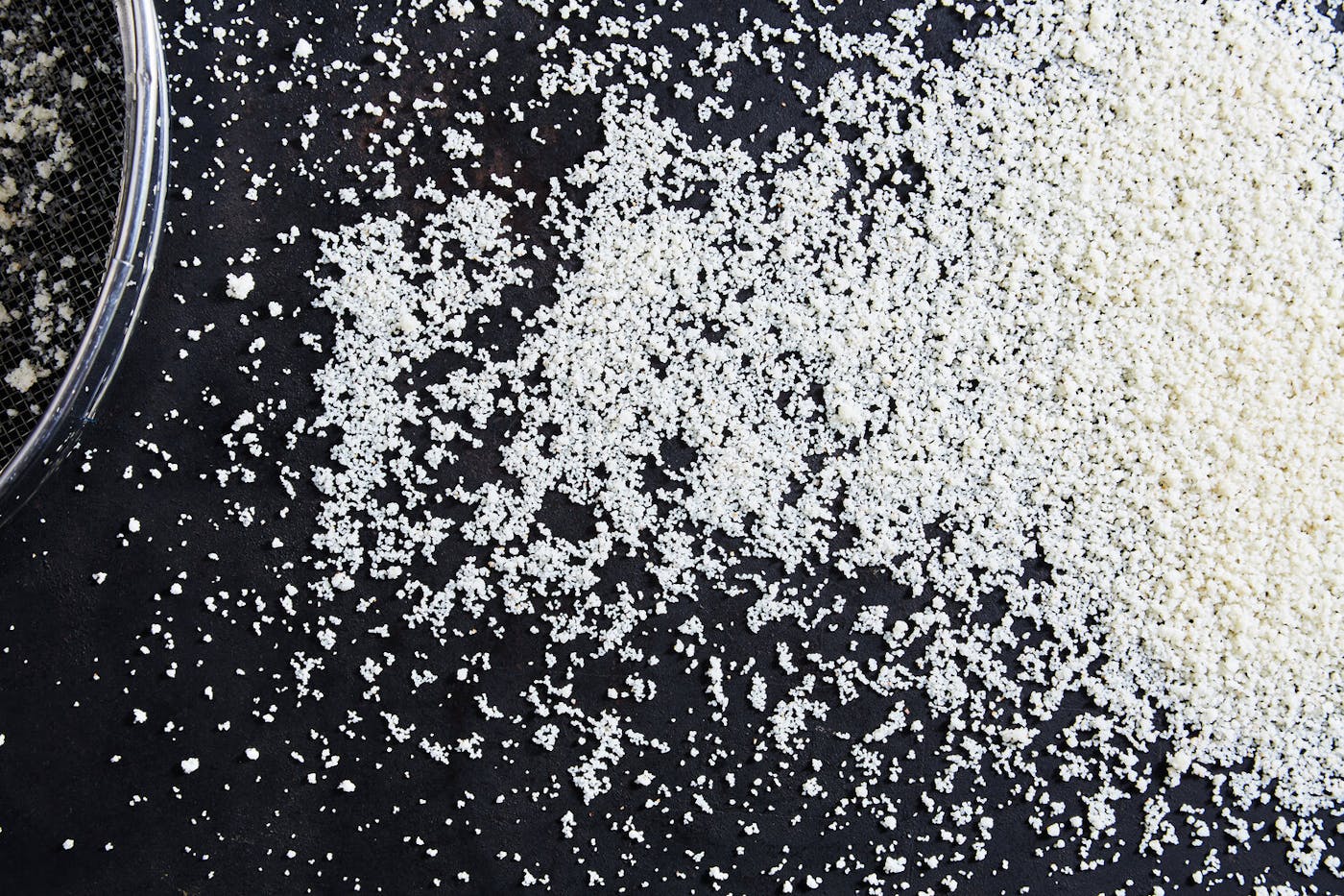 Freshly hand-rolled couscous spread onto black surface alongside sieve.