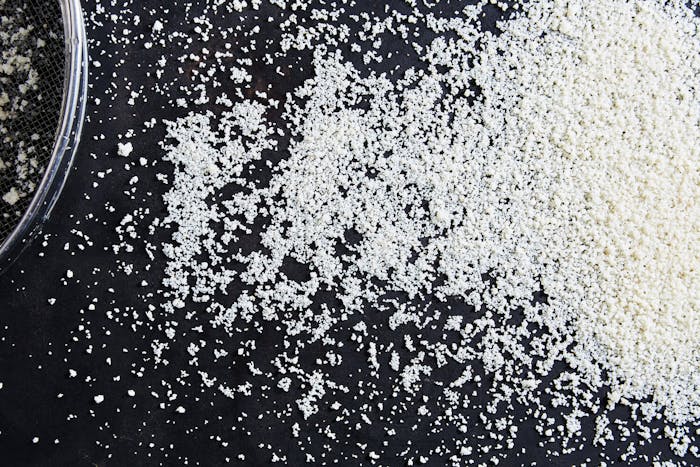 Freshly hand-rolled couscous spread onto black surface alongside sieve.