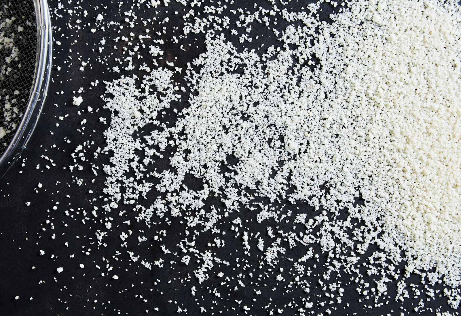 Freshly hand-rolled couscous spread onto black surface alongside sieve.