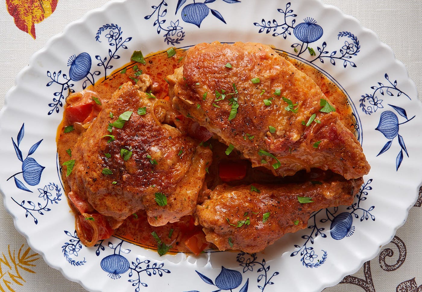 Chicken paprikas sprinkled with chopped parsley on blue-patterned plate, atop white tablecloth.