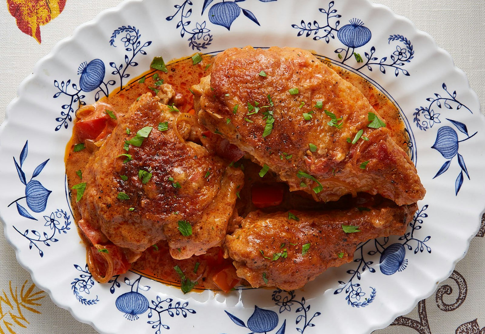 Chicken paprikas sprinkled with chopped parsley on blue-patterned plate, atop white tablecloth.